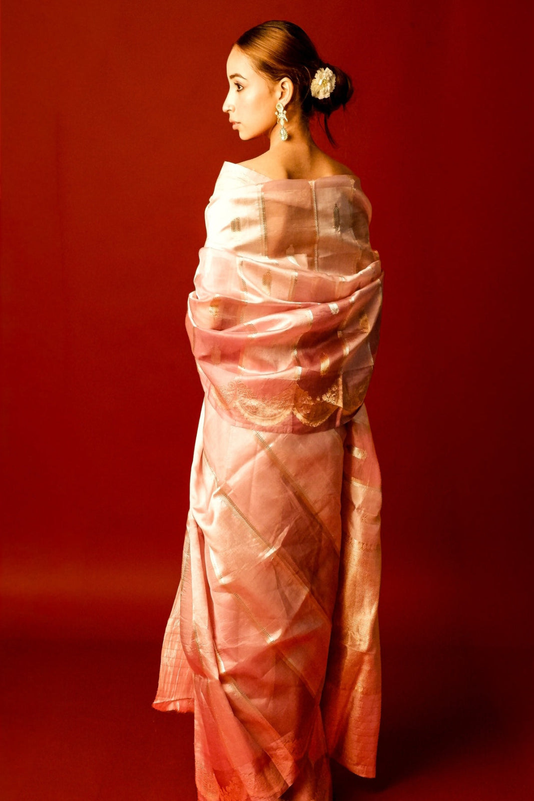A woman in profile wearing a draped light pink saree with subtle gold zari detailing, posed against a solid deep red studio backdrop.