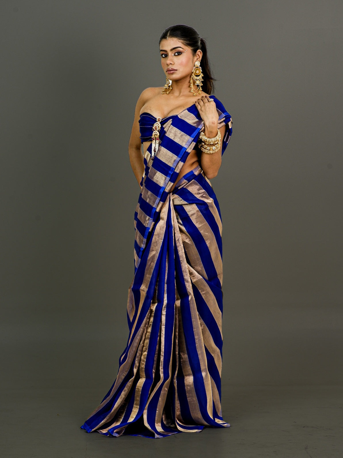 A woman modeling a royal blue and gold striped silk saree with a strapless sweetheart bodice, set against a solid dark gray studio background.