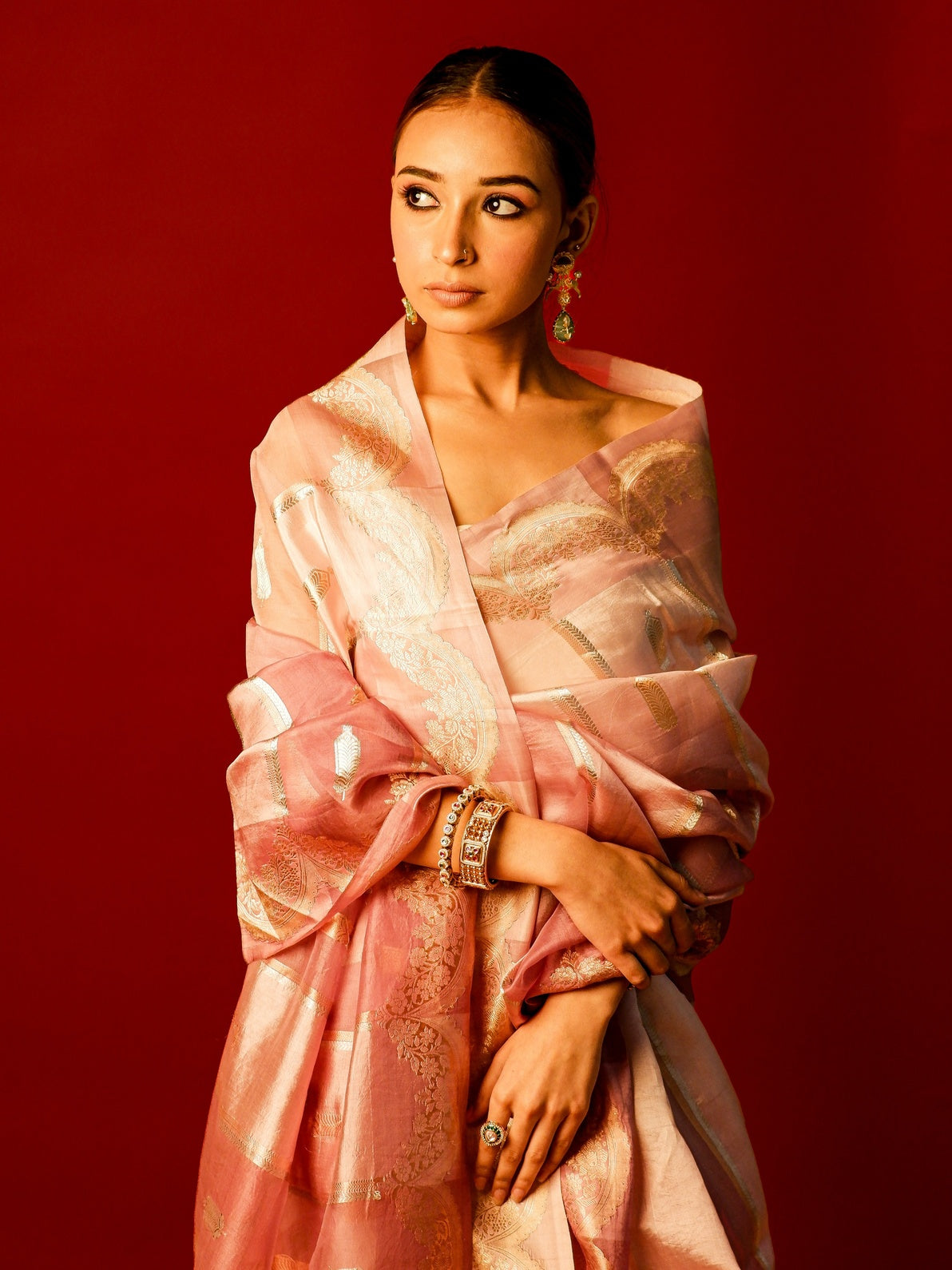 A woman wearing a shimmering pink and gold sari with ornate gold jewelry, posed against a solid deep red studio background.