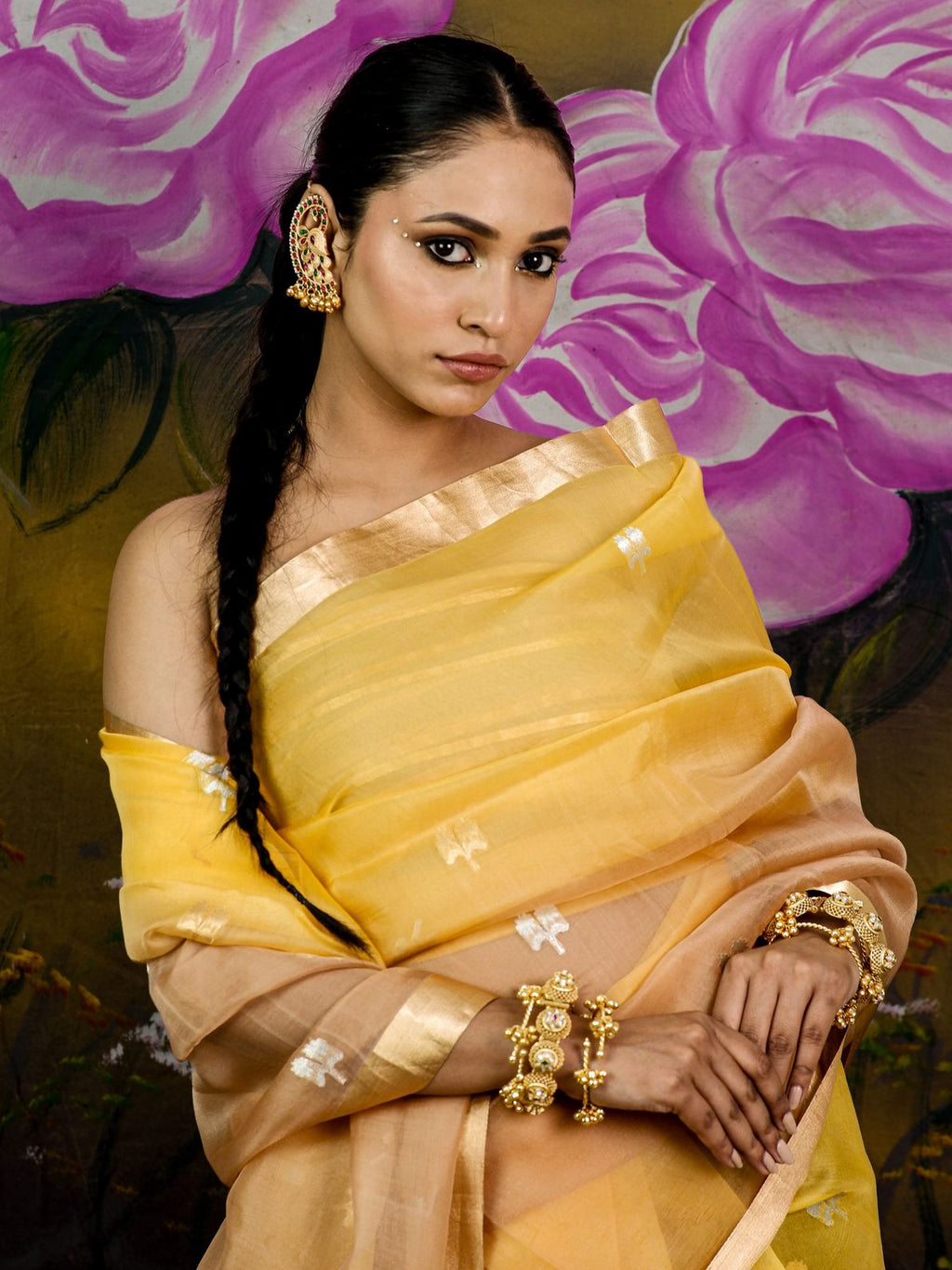 A woman wearing a yellow and beige saree with gold jewelry, posed against a painted background of large pink flowers.