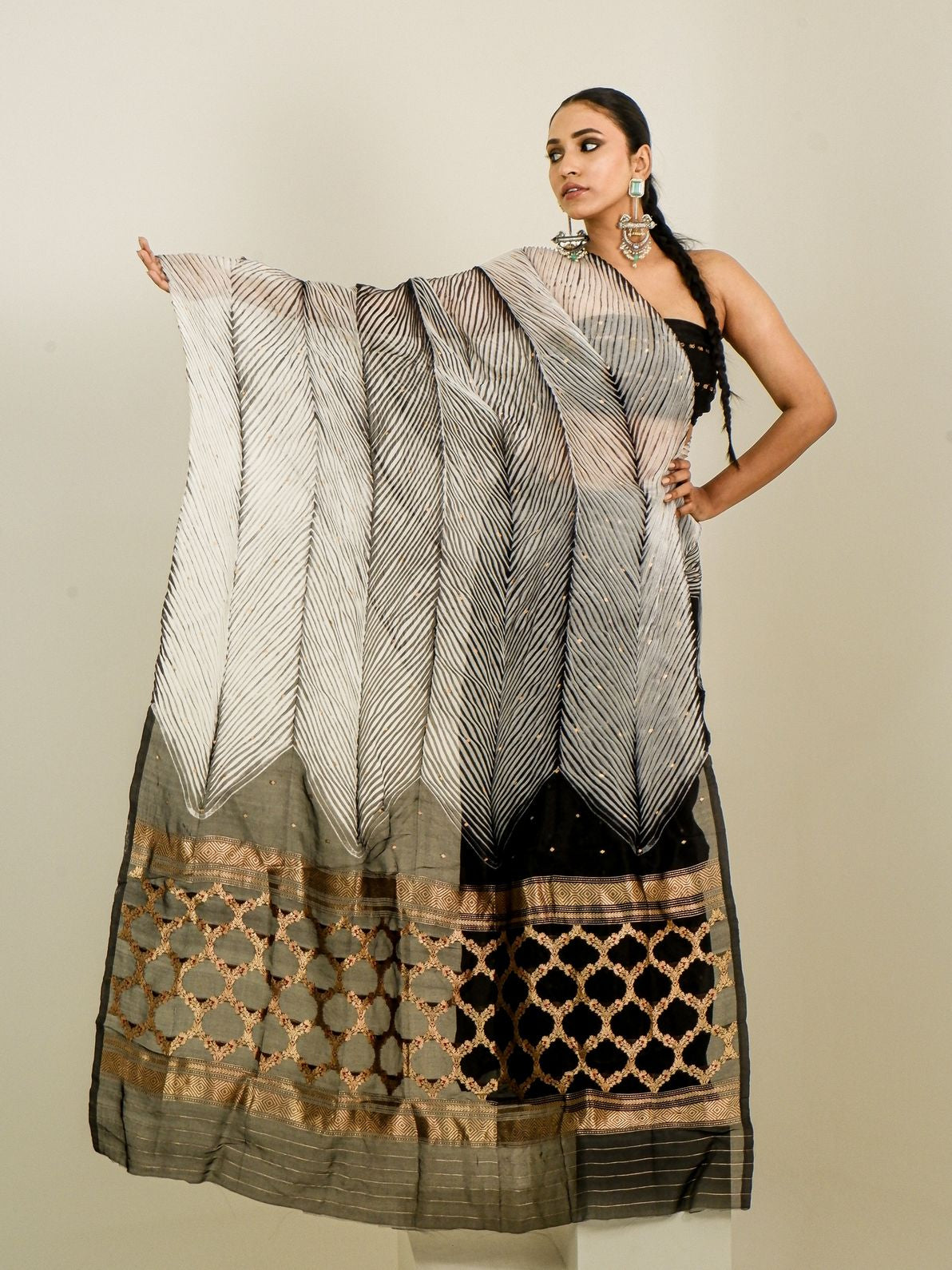 A woman models a traditional Indian saree with a black and white chevron-patterned body, a black and gold border, and a matching dupatta draped over her arm, set against a plain beige background.