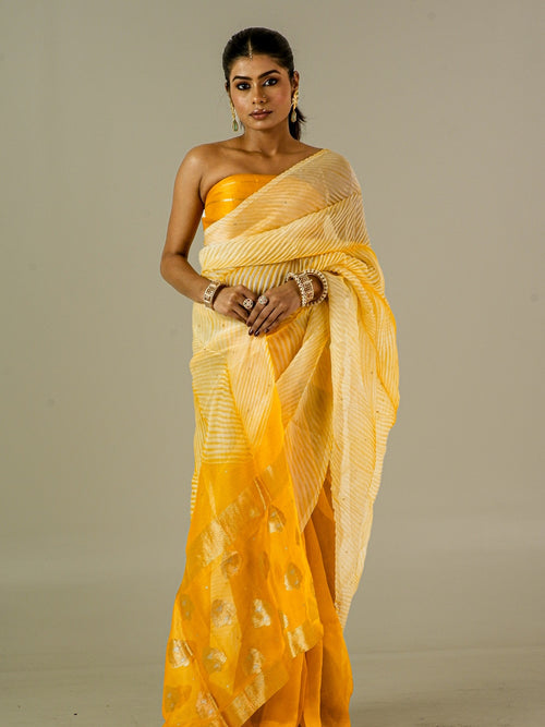 A woman modeling a yellow and cream striped saree with gold foil motifs, paired with a matching yellow strapless blouse, set against a plain light gray studio background.