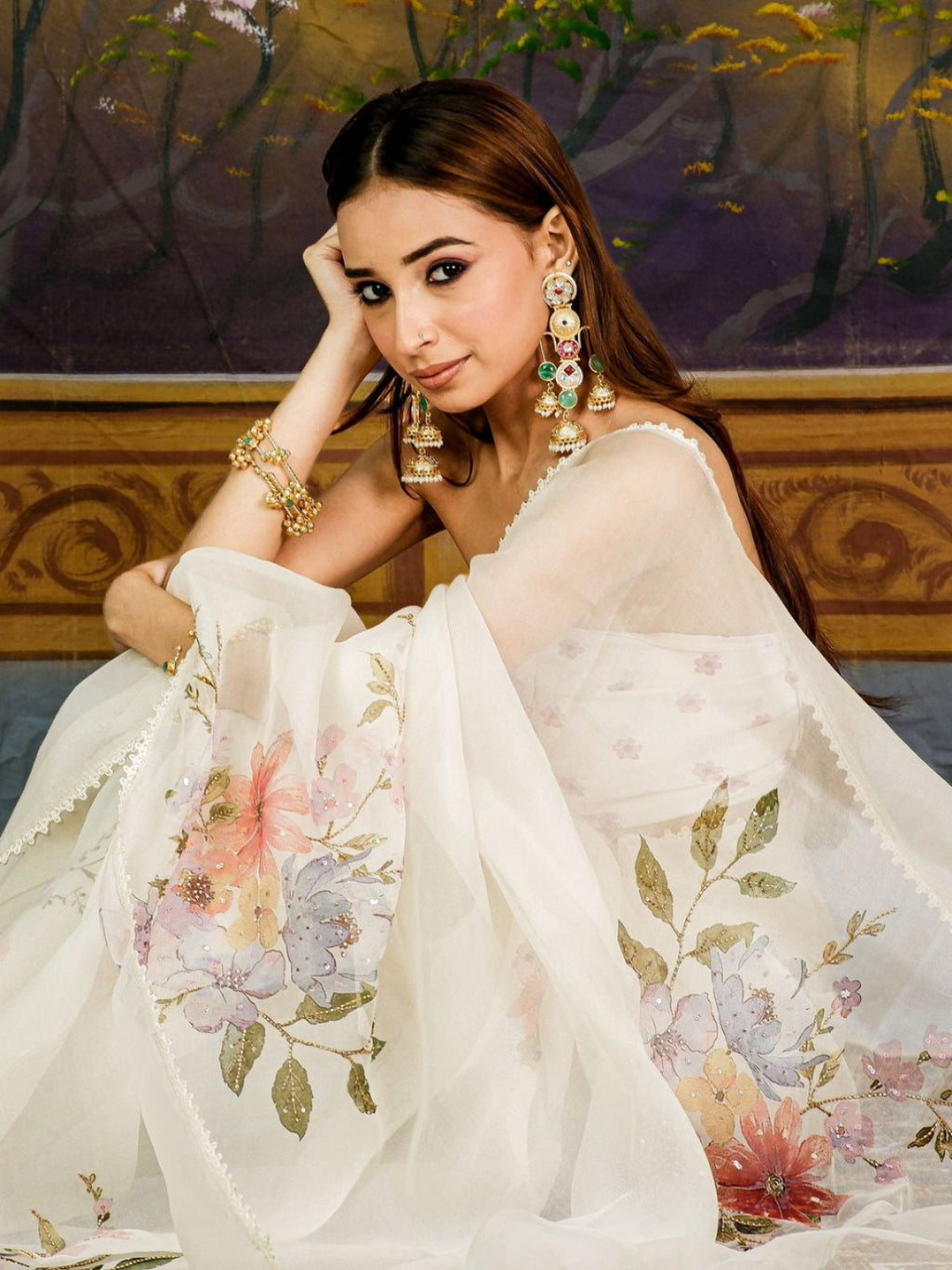 A woman wearing a white embroidered shawl or dupatta with pastel floral prints, styled with ornate gold jewelry, posed in front of a painted wooden backdrop.